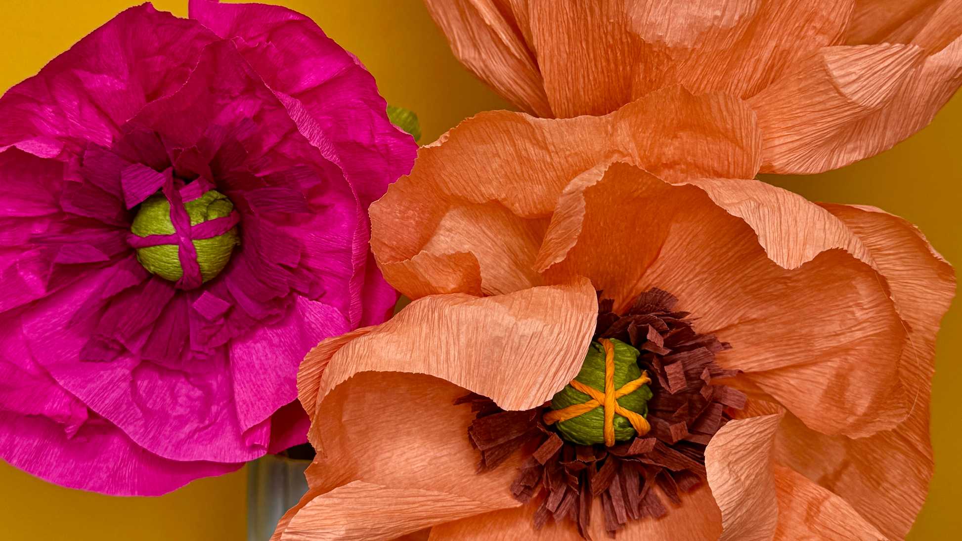 Two paper flowers, one pink and one peach, in a blue vase against a yellow background.